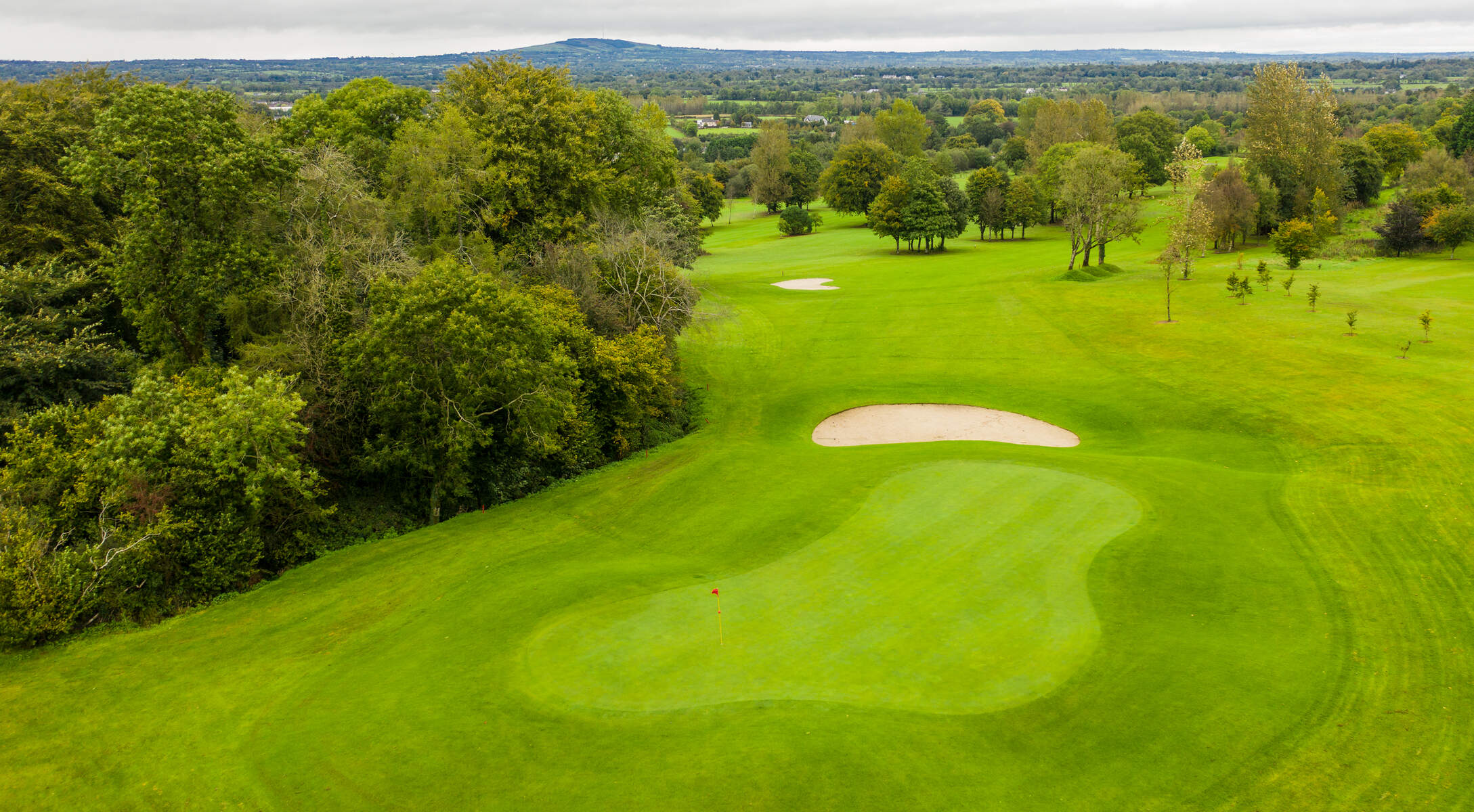 Scenic golf course with lush greenery, sand bunker, and distant hills under a cloudy sky.