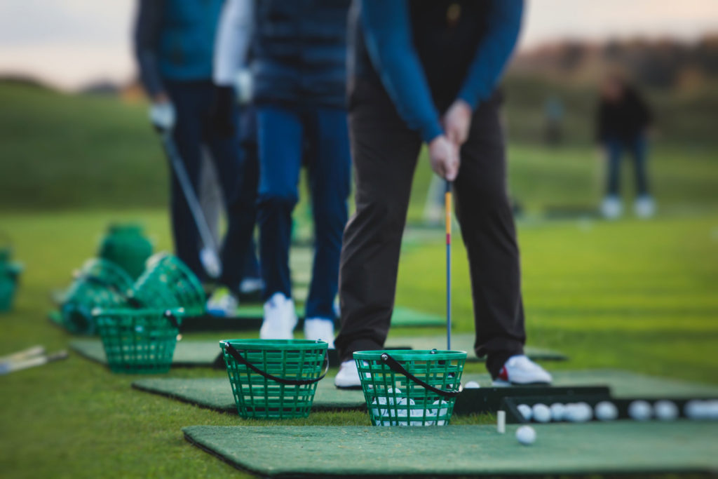 Golfer practicing swings on driving range with green baskets and scattered golf balls in focus.