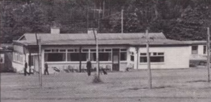 Vintage photo of a rural building with people walking outside, surrounded by trees.
