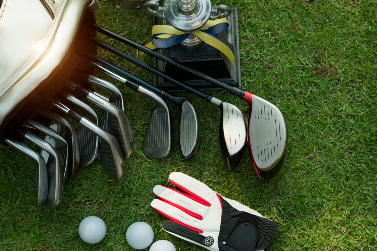 Set of golf clubs and glove on grass beside trophy and golf balls, ready for a tournament.