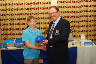 Boy receiving an award from an official at a ceremony, both smiling, with a table and patterned curtains in the background.