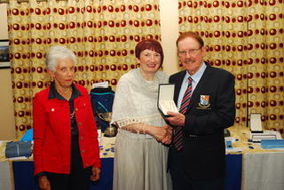 Three people at an award ceremony with a man holding a trophy. A table with gifts and curtains in the background.