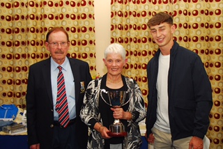 Three people at an award ceremony, with a woman holding a trophy, smiling in front of patterned curtains.