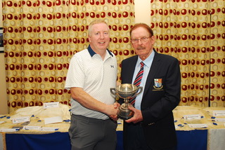 Two men holding a trophy at a ceremony, standing in front of a table and patterned curtains.