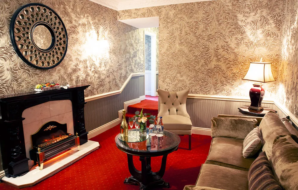 Elegant hotel lounge with vintage decor, plush sofa, ornate fireplace, and stylish mirror on patterned walls.